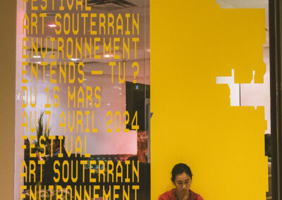 Une jeune femme, en train de dessiner, assise au pied d'une affiche en vitre du Festival Art Souterrain